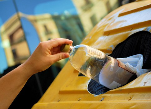 A person's hand, partially visible, is seen holding a clear plastic bottle over a yellow skip or container, positioned outdoors on a paved surface. The bottle appears to contain water or a clear liquid, and the person's fingers are grasping the neck of the bottle. The skip or container is made of metal with a smooth, glossy yellow finish, and has an open top showing a black interior. In the background, blurred building facades and reflections in nearby windows suggest an urban environment. The scene is well-lit, likely by natural daylight, emphasizing the reflective surfaces of the plastic bottle and the shiny yellow finish of the skip, which is part of a rubbish removal setup in a residential or commercial area possibly within Muswell Hill, north London. This image relates to waste management services provided by Commercial Waste Muswell Hill for efficient rubbish collection and disposal.