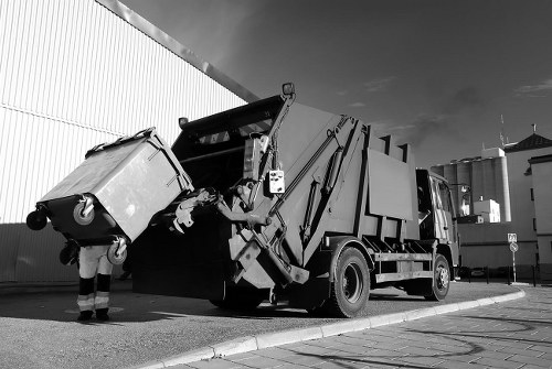 A commercial rubbish collection truck is positioned on a paved street near a large industrial building, with a worker in high-visibility clothing operating the hydraulic lifting mechanism. The truck has a black and white exterior with visible hydraulic arms extending towards a rear-compartment opening designated for waste containers. A large, wheeled bin made of durable plastic or metal, featuring a smooth surface and visible lid, is being lifted or emptied into the rear of the vehicle. The bin is positioned on the ground at an angle, with part of its body leaning toward the truck, suggesting an active loading process. In the background, there are additional industrial structures and a clear sky, indicating daytime. The setting appears typical of commercial waste collection areas, with clean, organized surroundings. This image reflects professional rubbish removal services relevant to businesses and properties in Muswell Hill or surrounding areas, such as those offered by Commercial Waste Muswell Hill.