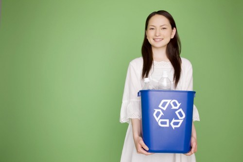 A young woman with long dark hair, smiling gently, stands outdoors against a solid light green background. She is dressed in a white, loose-fitting dress with lace details on the sleeves. In her hands, she holds a medium-sized blue recycling bin made of plastic, featuring a prominent white recycling symbol on the front. The bin appears clean and empty, with a simple, functional design suitable for household waste. The setting suggests a focus on environmental consciousness and waste management, aligning with services provided by Commercial Waste Muswell Hill in North London. The image emphasizes the importance of proper waste segregation and recycling practices, supporting local rubbish removal and recycling initiatives in the Muswell Hill area. The bright natural lighting enhances the clarity of the scene, highlighting the vibrant blue of the bin and the cheerful expression of the woman, conveying a message of responsible waste disposal and sustainability.