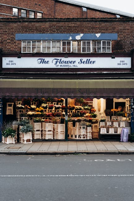 The image depicts the storefront of 'The Flower Seller of Muswell Hill,' a small flower shop situated beneath a dark brown awning with the shop's name printed in white. The shopfront is filled with an