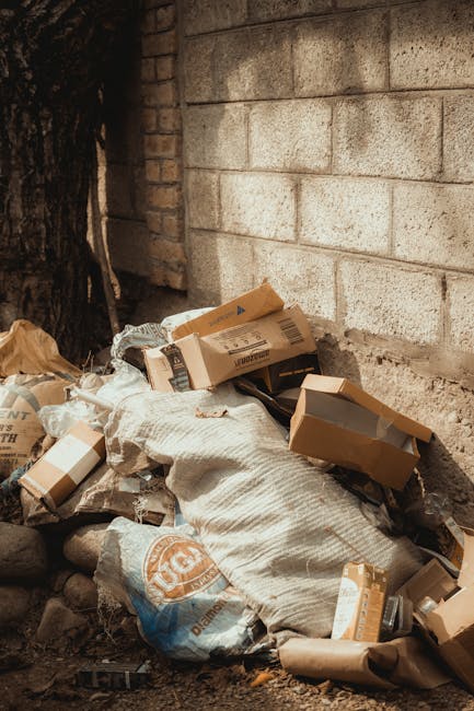 A pile of discarded cardboard boxes, some flattened and others still partially intact, are haphazardly stacked against a rough brick wall and a tree trunk in an outdoor setting. The boxes vary in size