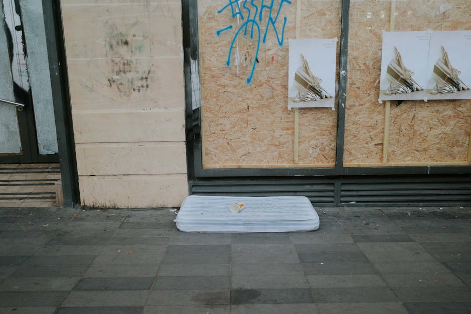 A discarded white foam mattress is lying flat on a paved sidewalk next to a building wall. The mattress appears to be used, with some creases and dirt marks visible on its surface. A small, crumpled p